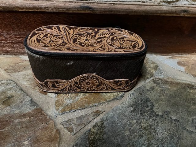 A black and brown leather Marshal Hand Tooled Make-Up Kit sitting on a stone floor.